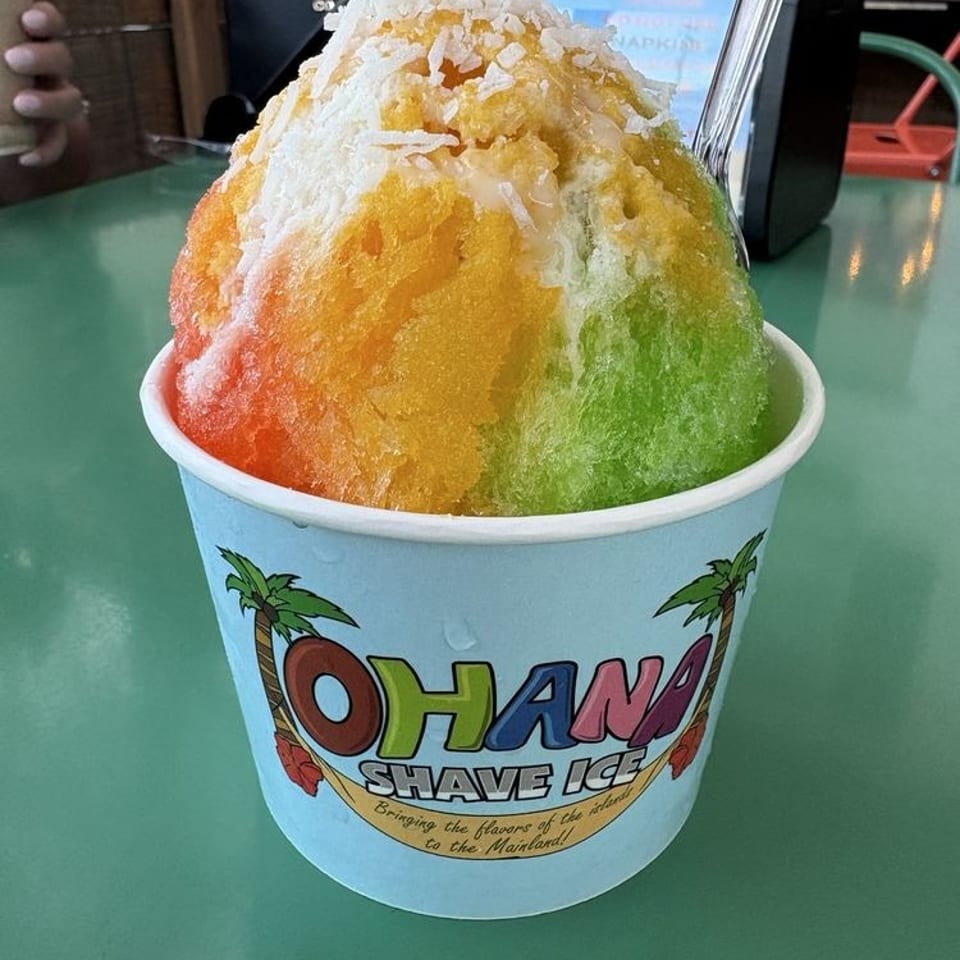 Colorful rainbow shaved ice topped with coconut flakes in an Ohana Shave Ice cup on a green table.