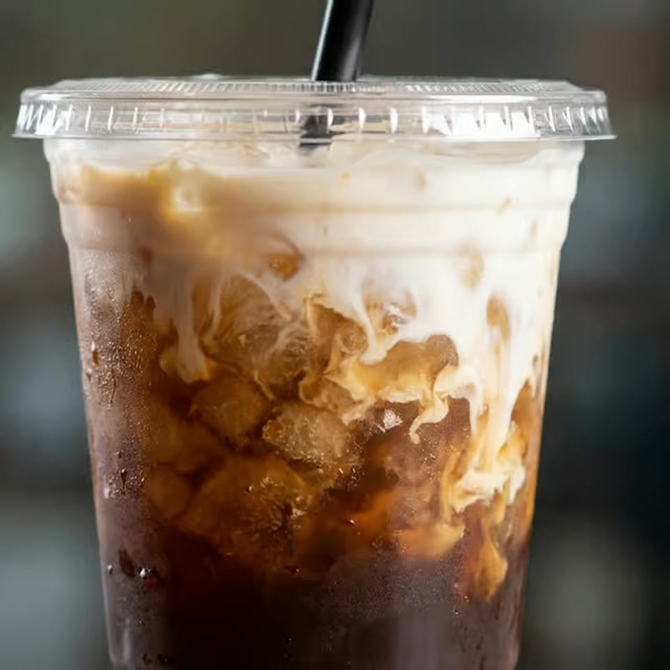 Close-up of iced coffee with creamy foam swirling through the drink in a clear plastic cup with straw.