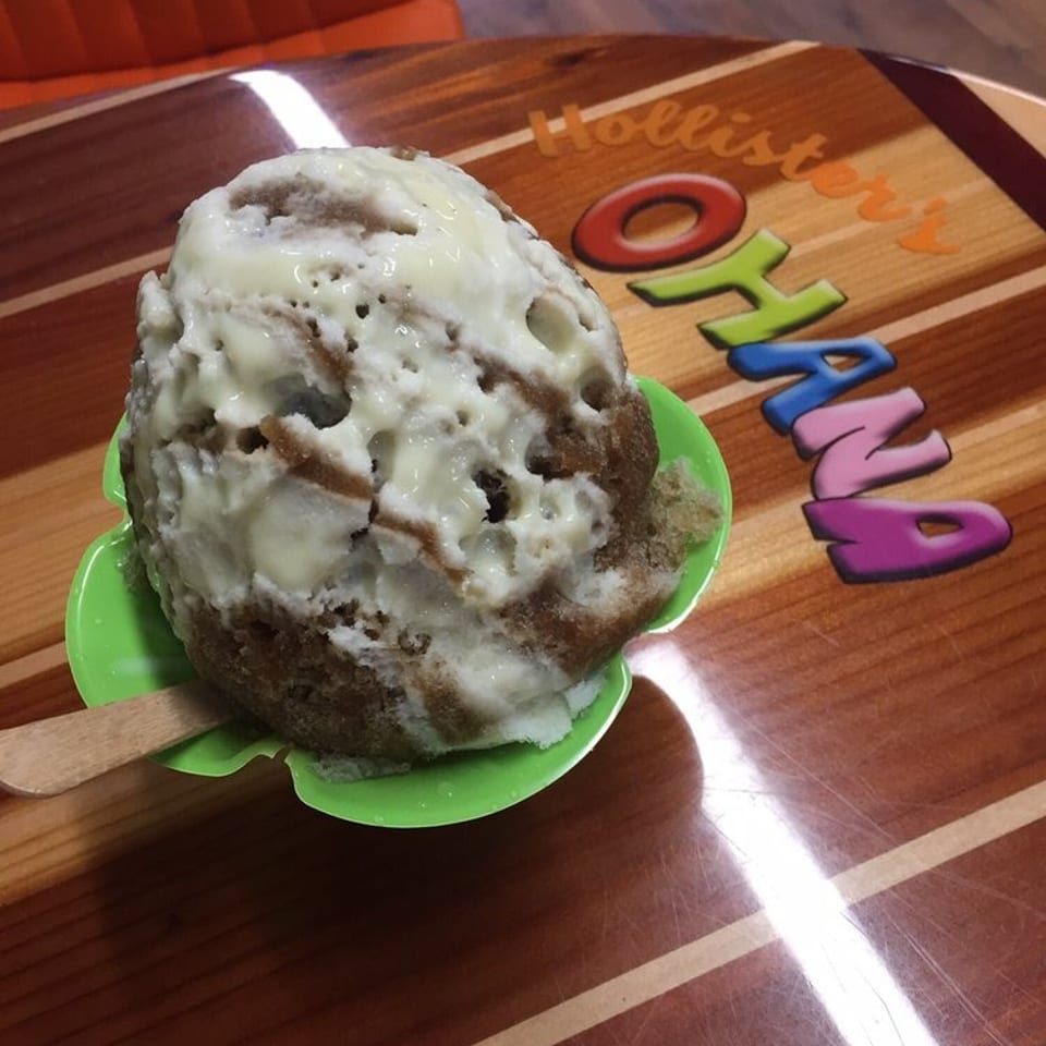 Large shaved ice dessert drizzled with white topping in a green bowl on a colorful wooden table.