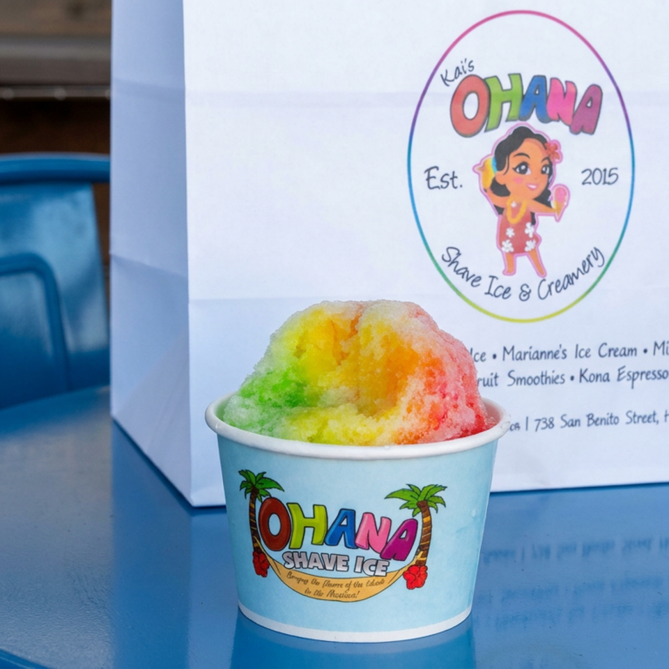Rainbow-colored shaved ice in an Ohana Shave Ice cup sits on a blue table beside a branded menu sign.