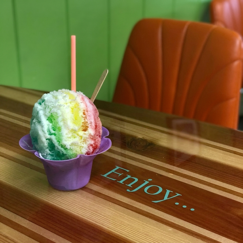 Colorful shaved ice in a purple cup with a straw on a wooden table, orange seating and green walls behind.