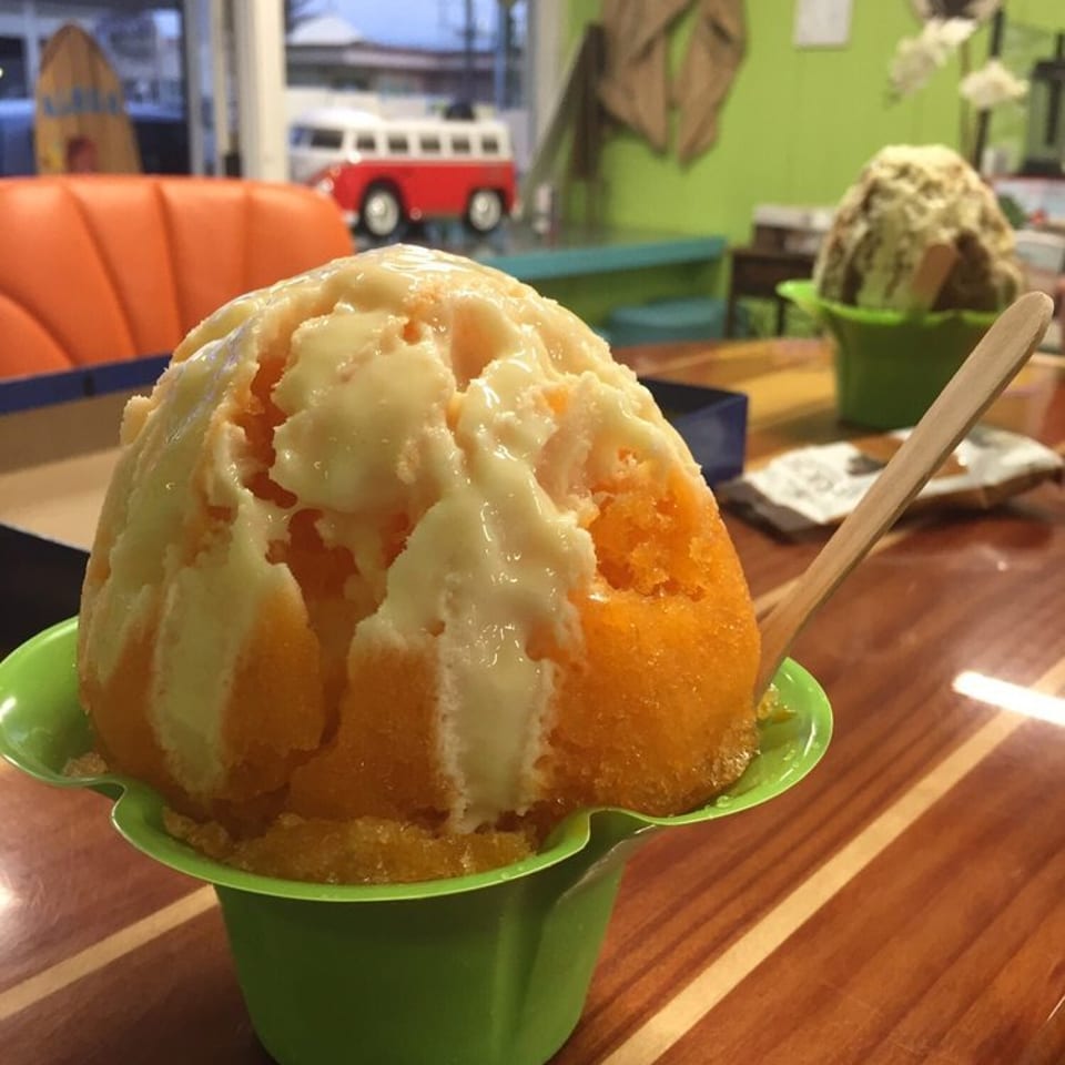 Orange shaved ice topped with creamy white drizzle in a green cup, served on a wooden table inside a colorful tropical-themed restaurant.