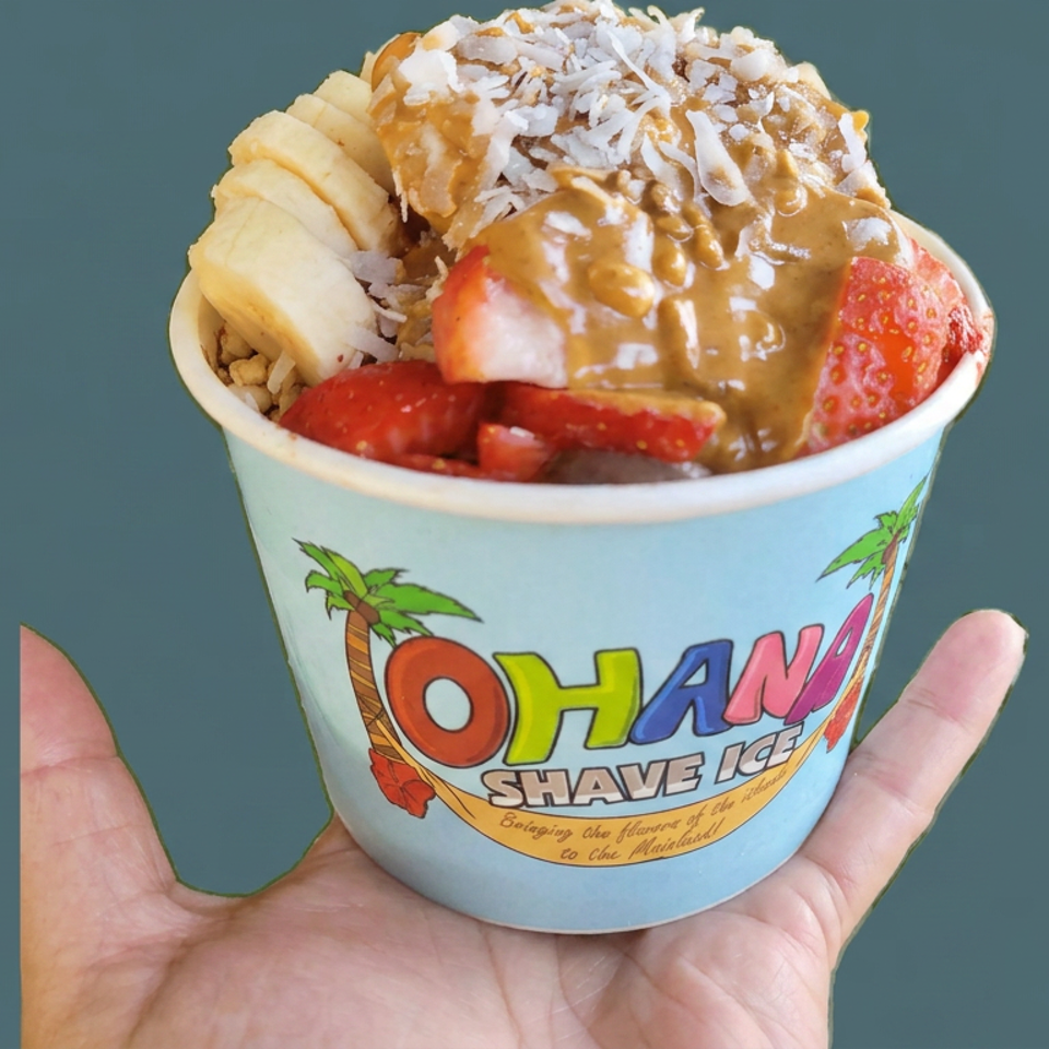 Hand holding an Ohana Shave Ice cup topped with bananas, strawberries, coconut, and brown syrup.