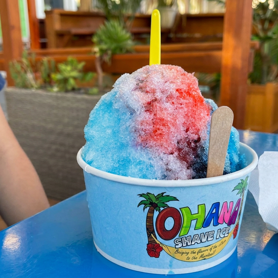 Colorful blue, pink, and red shaved ice in a branded cup with a wooden spoon, served outdoors at a tropical restaurant.